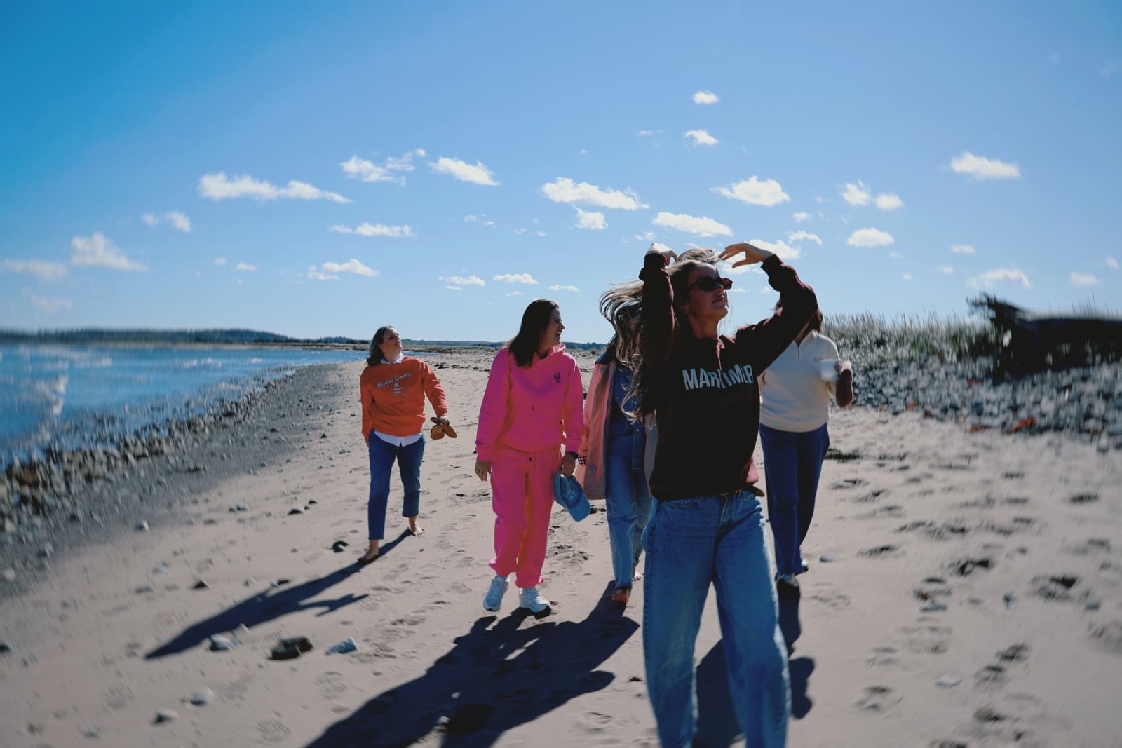 Friends in colourful sweatshirts walking together on a sandy beach beside calm blue water