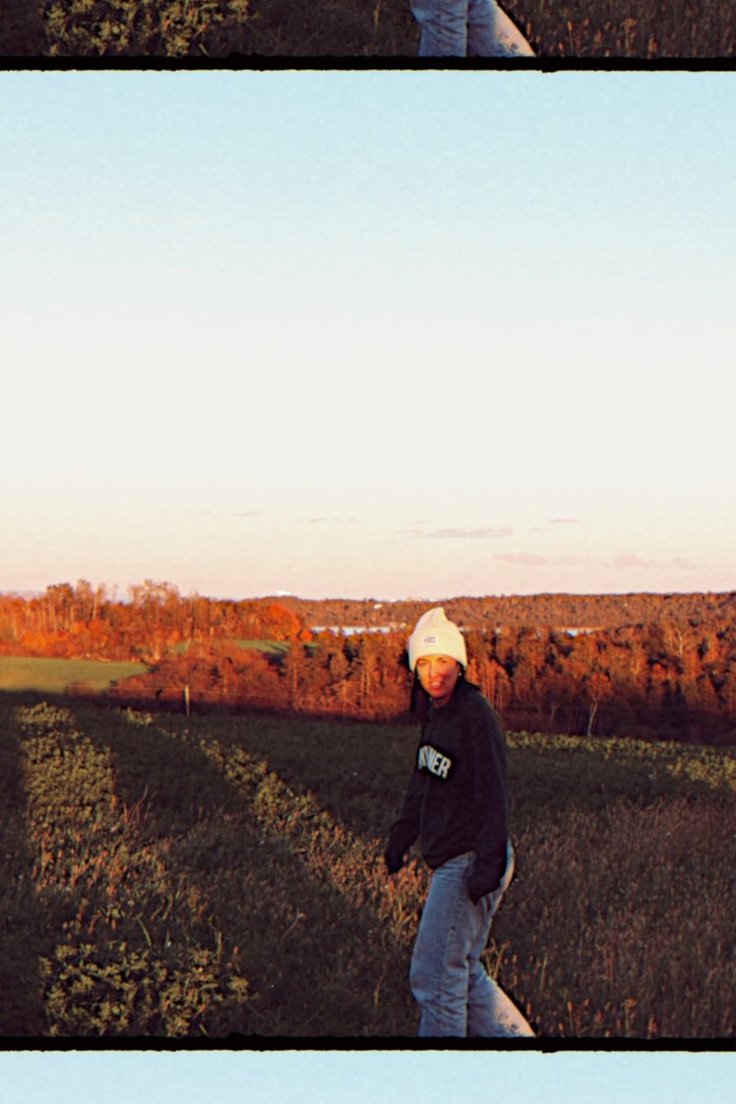 Film-bordered portrait of a woman in a cream beanie and dark sweatshirt walking through an autumn field
